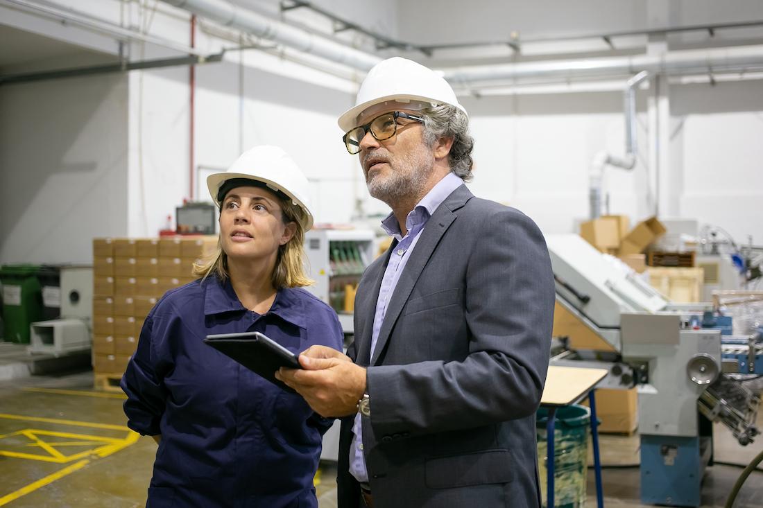 Consultor da Genesia com tablet e responsável de produção, ambos de capacete, avaliam uma unidade fabril — acompanhamento em financiamento de inovação e I&D.