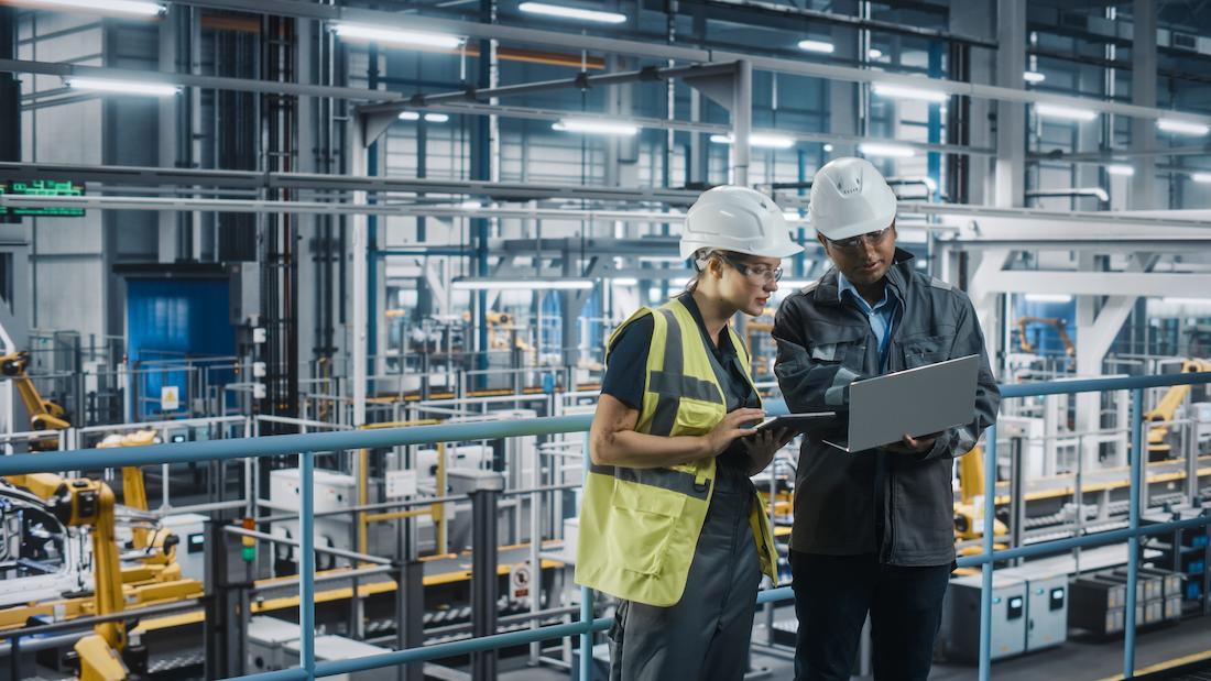 Dois engenheiros com equipamento de proteção analisam dados num portátil numa fábrica automatizada — inovação e I&D industrial.